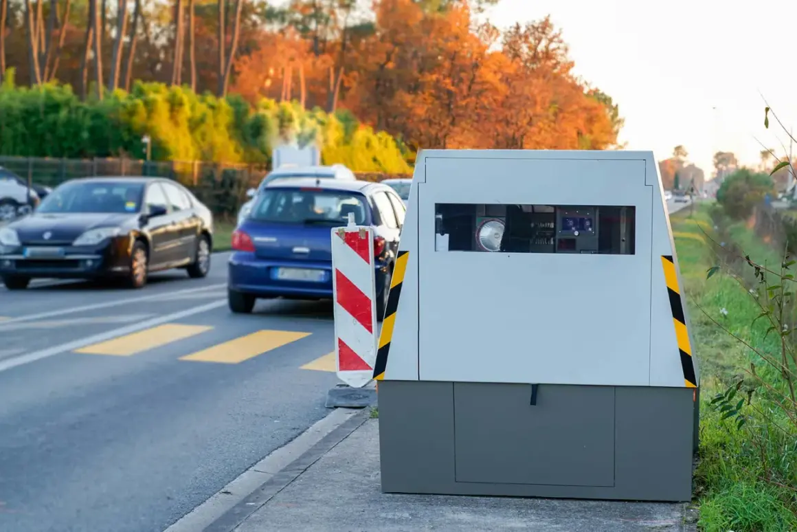 Un radar de chantier surveille la circulation sur une route bordée d'arbres aux couleurs d'automne. Des voitures passent devant.