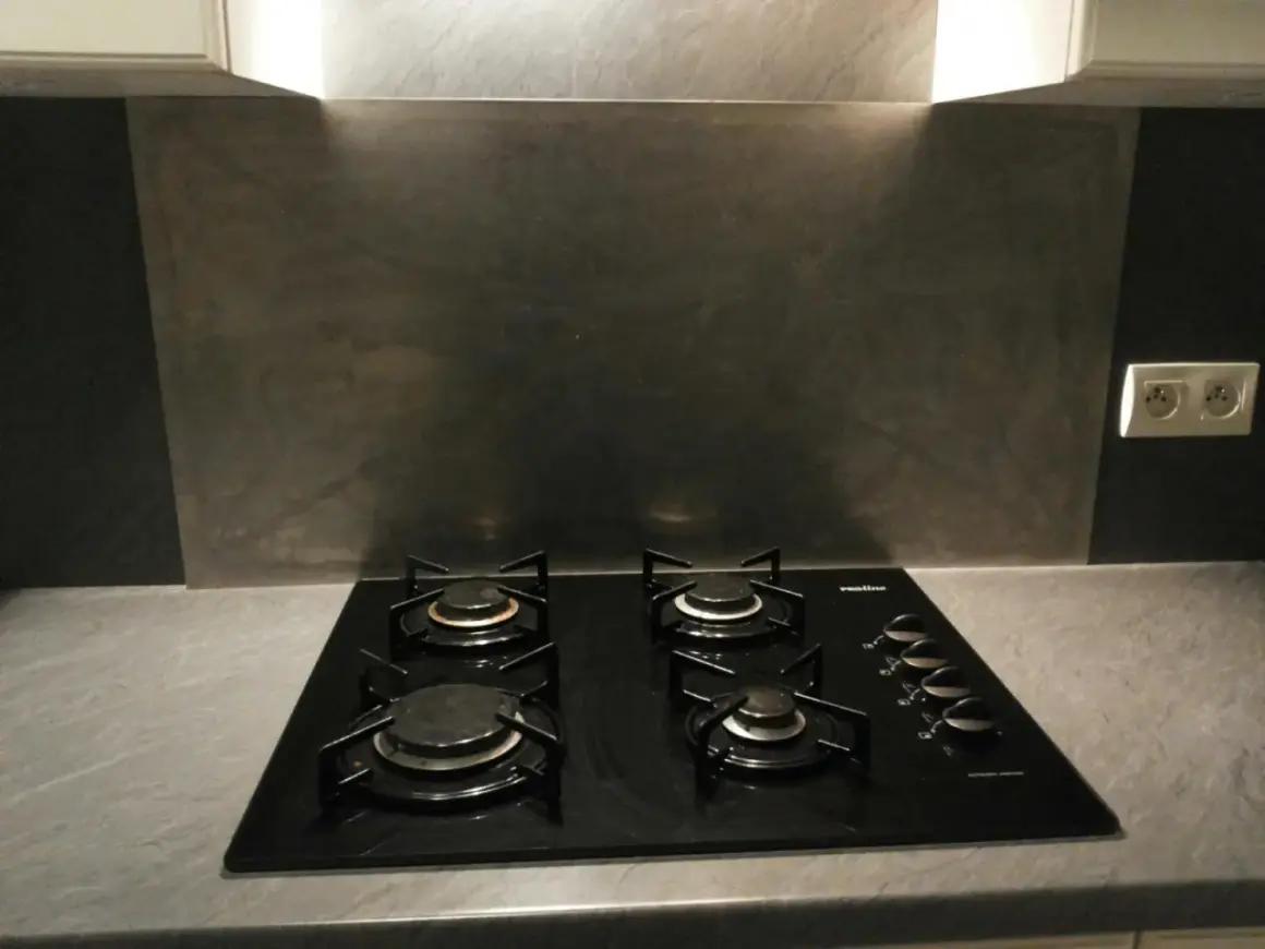 Plaque de cuisson noire avec quatre feux, devant un mur de crédence gris métallisé.