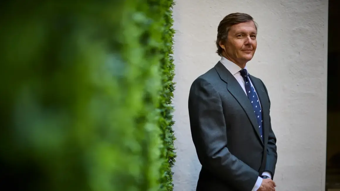 Cayetano de Borbón-Dos Sicilias, con traje gris y corbata azul, posa con una sonrisa ante una pared blanca y vegetación verde.