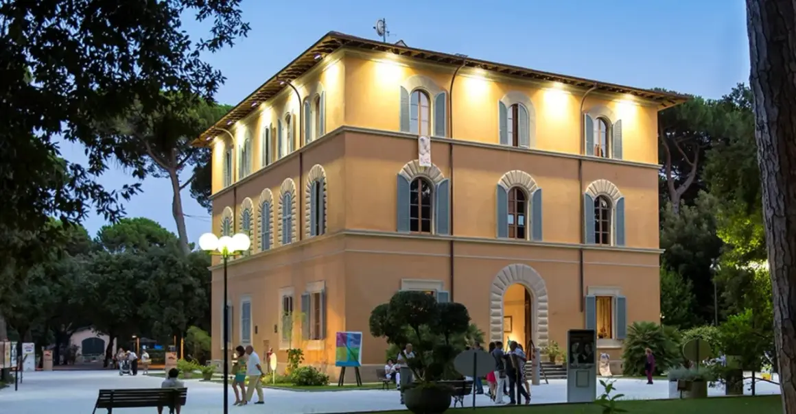 Edificio giallo illuminato nel parco della Versiliana, con persone che passeggiano e un'opera d'arte esposta.