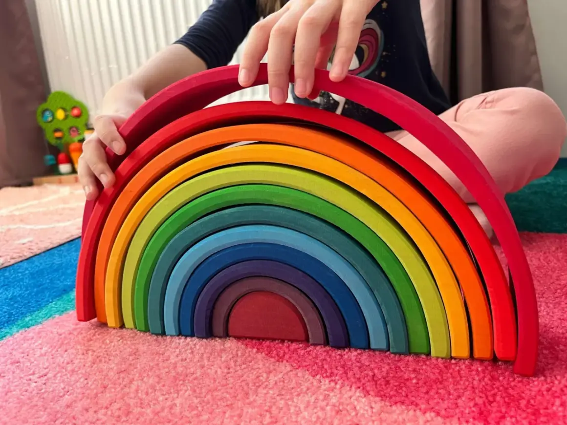 Dziecko bawi się tęczowym łukiem, jedną z popularnych zabawek, układając kolorowe drewniane elementy na dywanie.