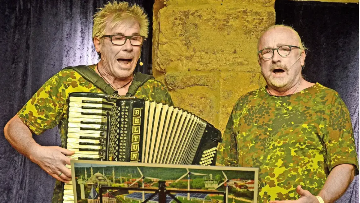 Zwei Männer im Tarnanzug singen und spielen Akkordeon beim **Kabarett der Galgenstricke**. Ein Bild mit Stadtansicht und Windrädern liegt vorn.
