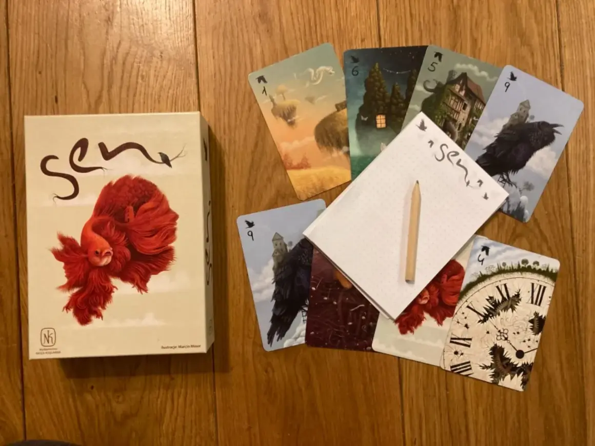 Pudełko gry karcianej "Sen" i rozłożone karty z surrealistycznymi ilustracjami, jak latające wyspy i mroczne zamki.