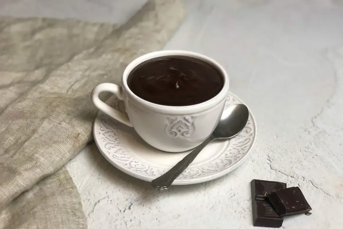Una tazza di cioccolata calda cremosa, perfetta per imparare come fare una buona cioccolata calda. Accanto, due quadratini di cioccolato fondente e un cucchiaino.