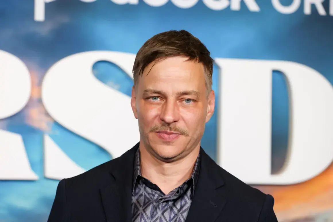 Tom Wlaschiha, mit einem Schnurrbart und blauen Augen, posiert vor einem Hintergrund mit großen weißen Buchstaben. Er denkt vielleicht an seine tom wlaschiha kinder.
