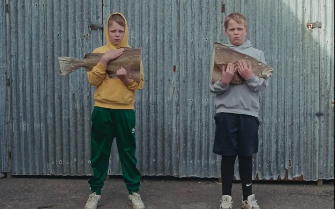 Två barn, som påminner om Sverrir Gudnason som barn, håller varsin fisk framför en plåtvägg.