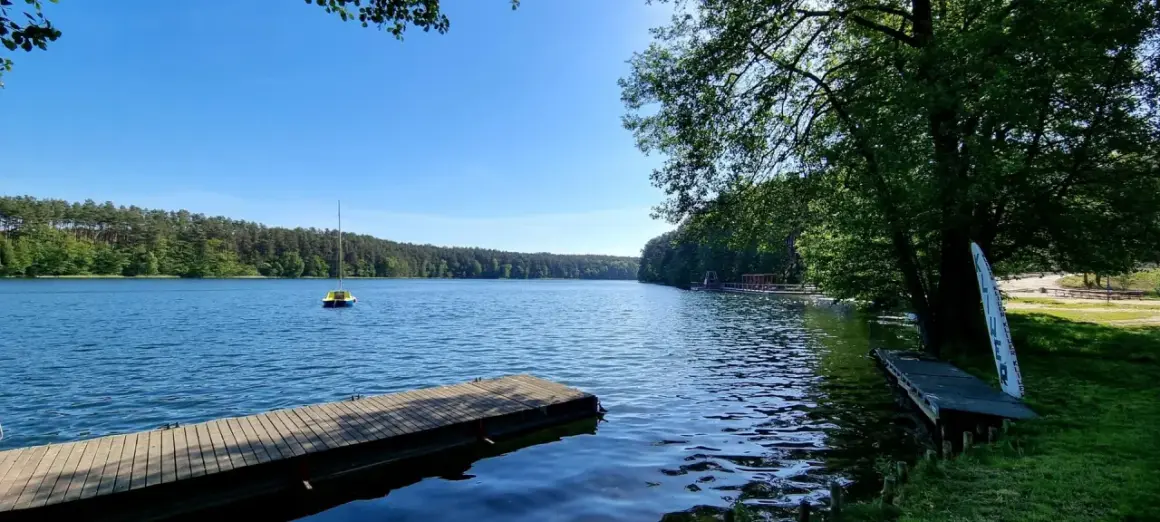 Drewniane pomosty nad spokojnym jeziorem, z małą żółtą łódką na wodzie i lasem w tle. Słońce odbija się od powierzchni, tworząc wrażenie, jakby jezioro było "spalone" blaskiem.