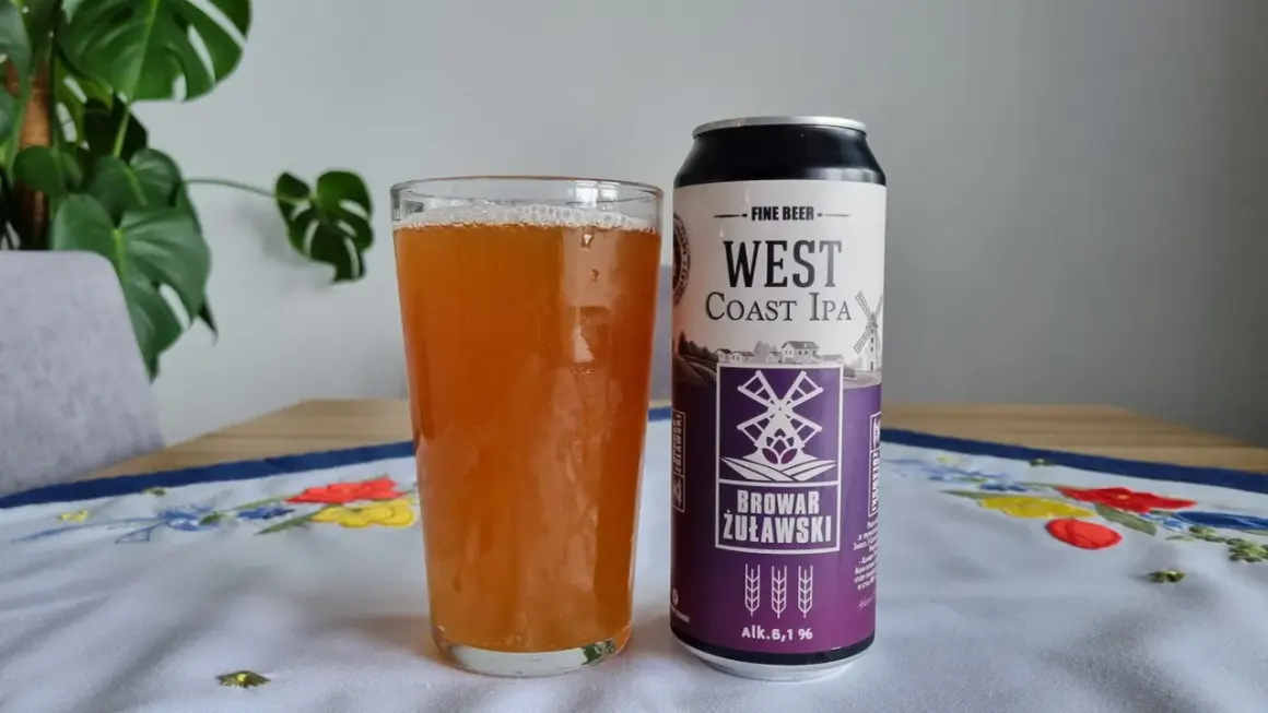 Szklanka piwa West Coast IPA obok puszki Browaru Żuławski. Na obrusie haftowane kwiaty.
