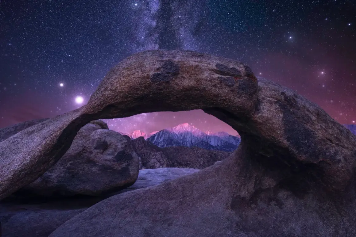 A natural rock arch frames a majestic mountain range under a breathtaking starry sky.