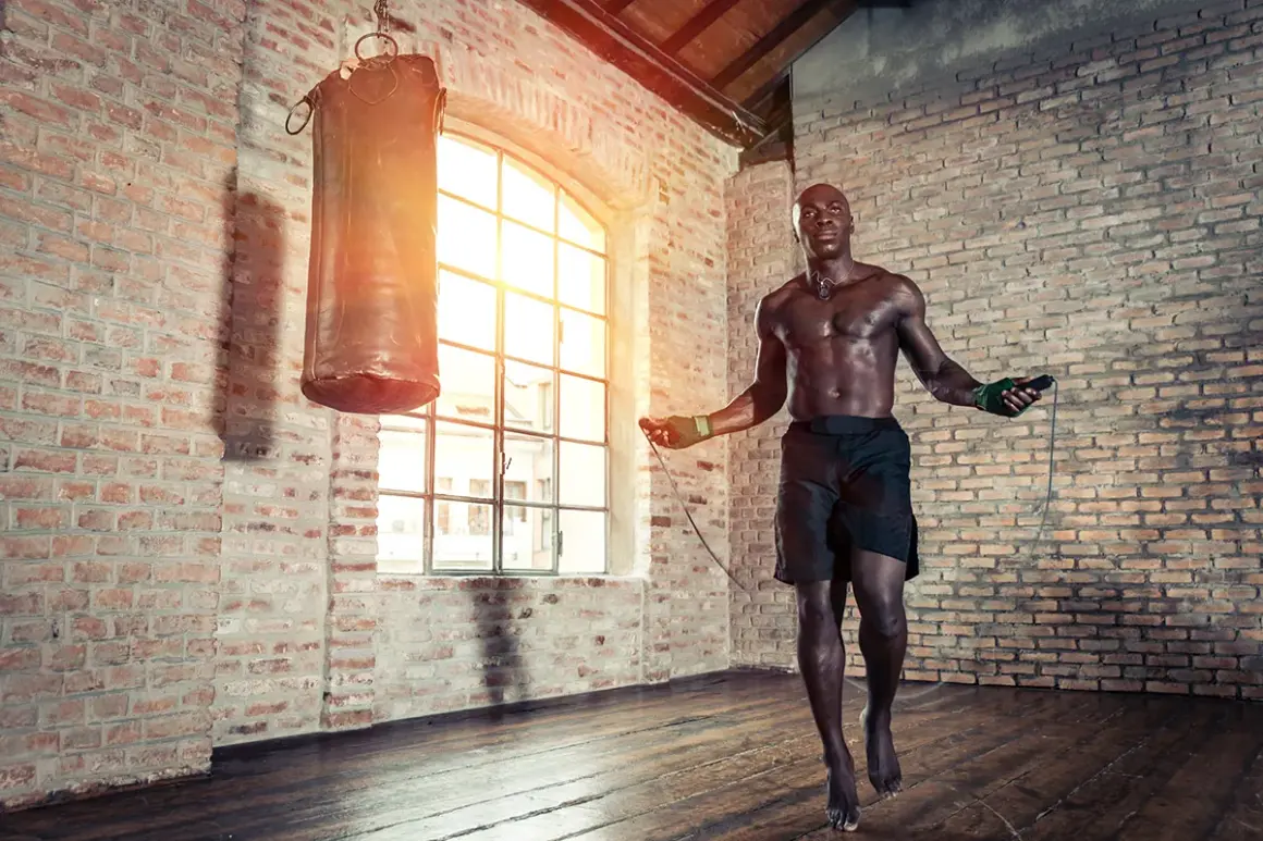 A muscular boxer practices how to jump rope like a pro in a brick gym, with a punching bag nearby.
