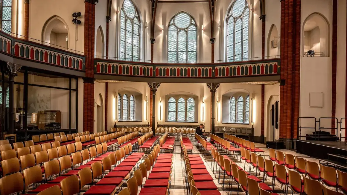 Innenraum einer epo church mit vielen roten Stuhlreihen und großen Bogenfenstern. Ein Mann sitzt allein.