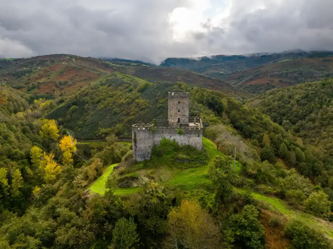 El **castillo de Doiras** se alza majestuoso sobre una colina verde, rodeado de montañas boscosas y un cielo nublado.