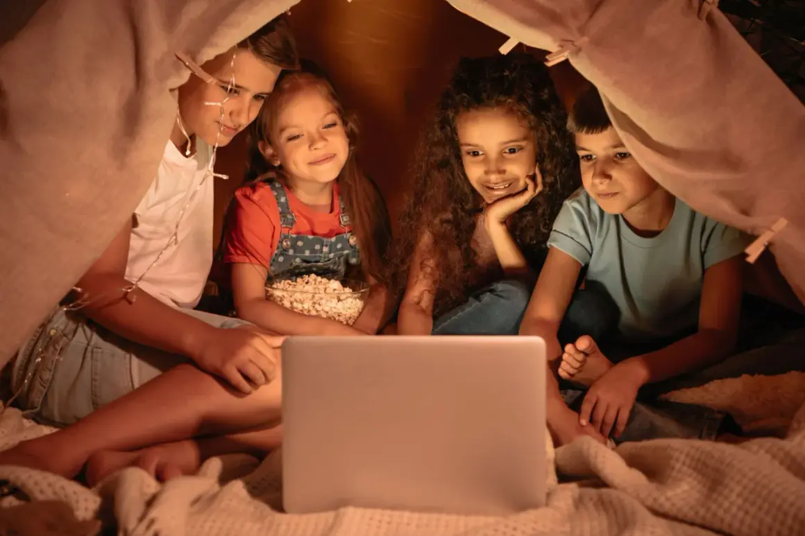 Quattro bambini guardano un film per ragazzi di 12 anni su un laptop, accoccolati in una tenda con luci scintillanti e popcorn.