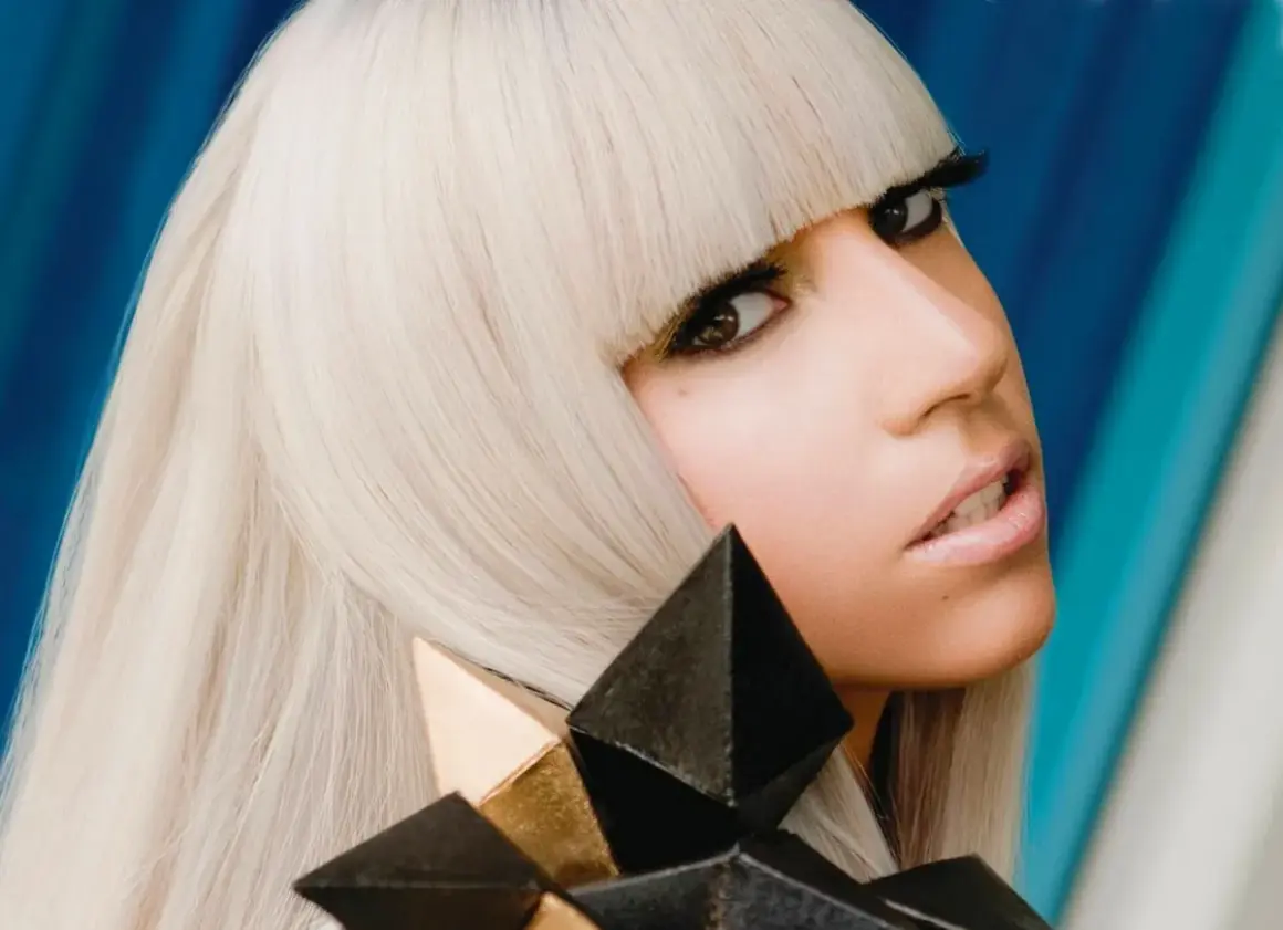 Lady Gaga, con capelli biondo platino e un look audace, cattura l'essenza del suo **inizio carriera** con un'espressione intensa e un accessorio geometrico.