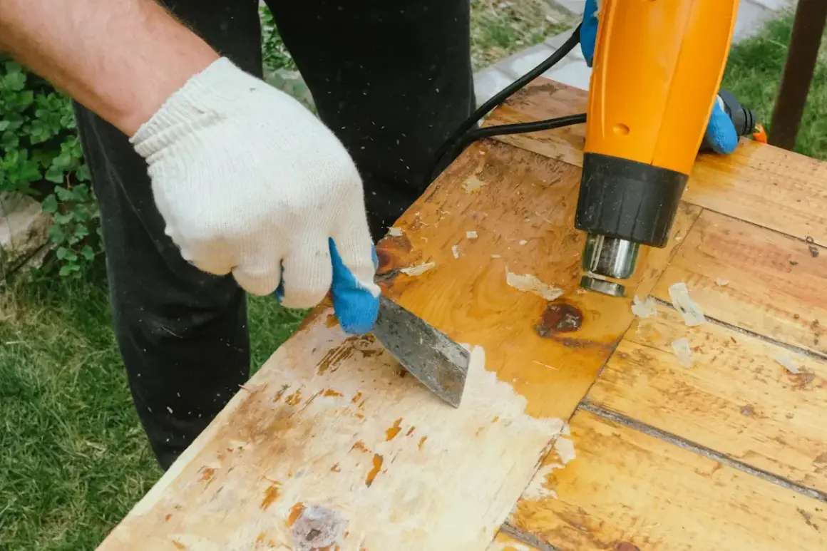Main tenant un décapeur thermique et une spatule pour retirer la colle sur bois vernis.