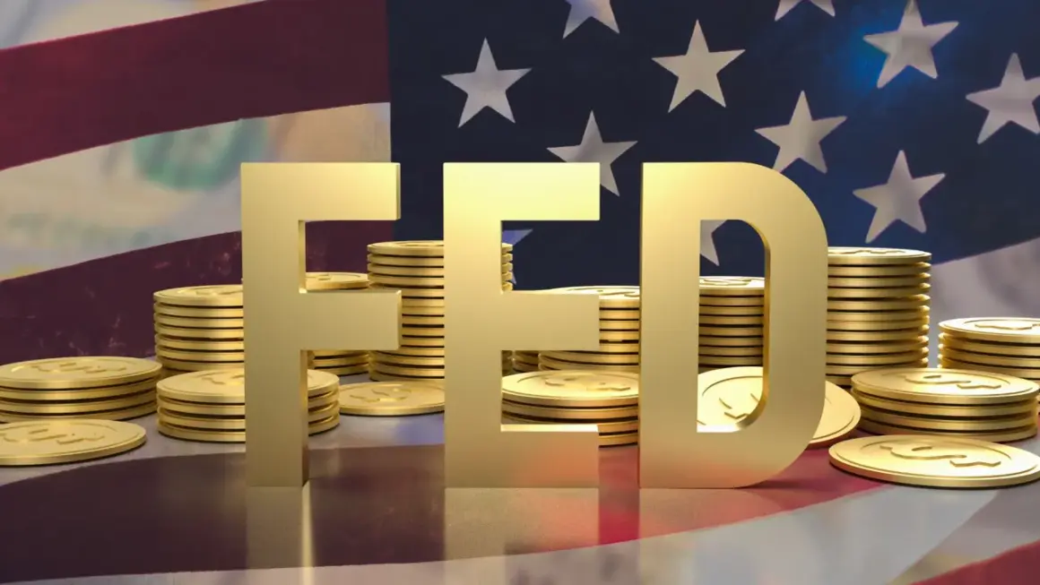 Złote litery "FED" na tle flagi USA i stosów monet. FOMC to komitet, który decyduje o polityce pieniężnej.