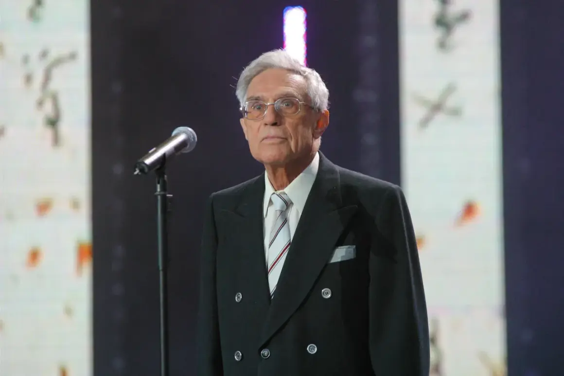 Starszy pan w okularach, Lucjan Szołajski, w czarnym garniturze i pasiastym krawacie stoi przy mikrofonie na tle sceny.