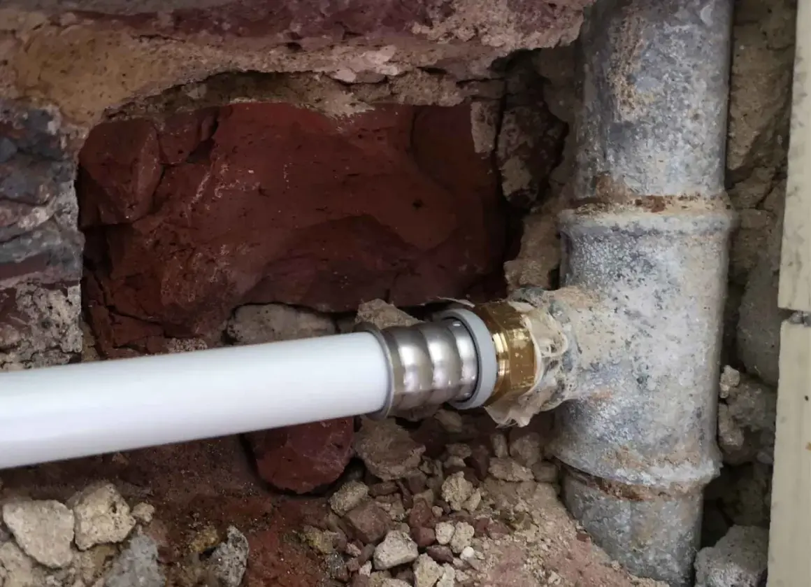 Un nouveau tuyau blanc est raccordé à un ancien tuyau de plomb pour l'eau. La maçonnerie est visible autour des canalisations.