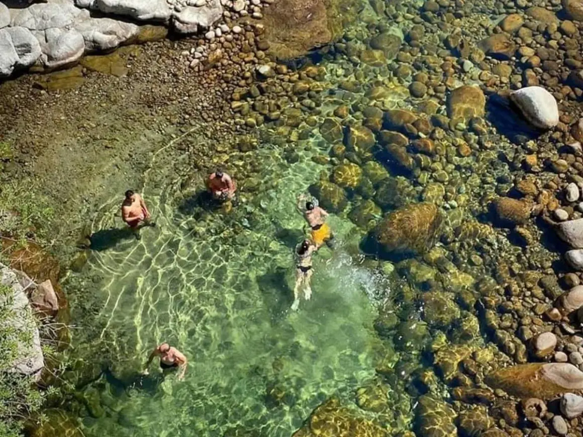 Gente se baña en un río cristalino con fondo de piedras, cerca de un **ponton de la oliva**.