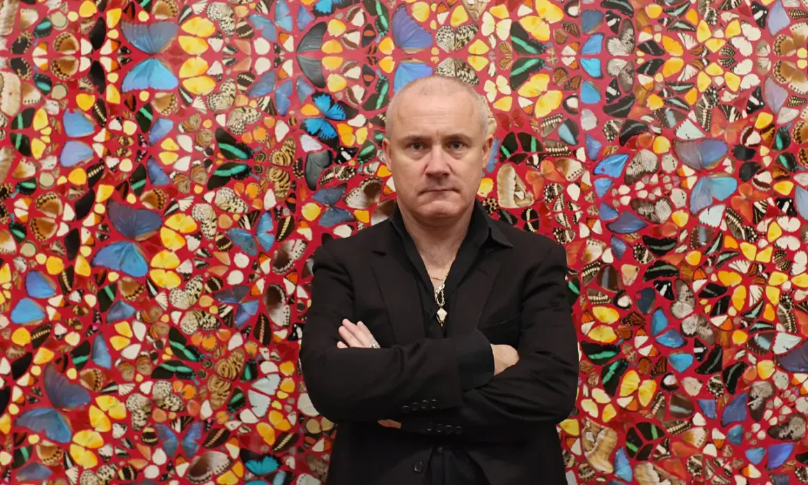 Damien Hirst stoi przed ścianą ozdobioną jego dziełami z motyli.