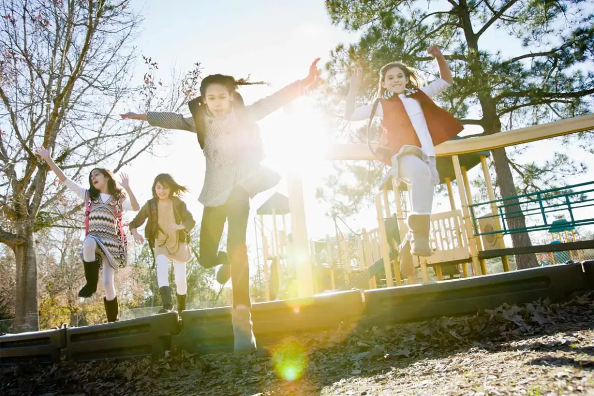 Vier fröhliche Kinder springen ausgelassen auf einem Spielplatz. Sie lernen in Bewegung, die Sonne scheint, und ihre Arme sind in die Luft gestreckt.