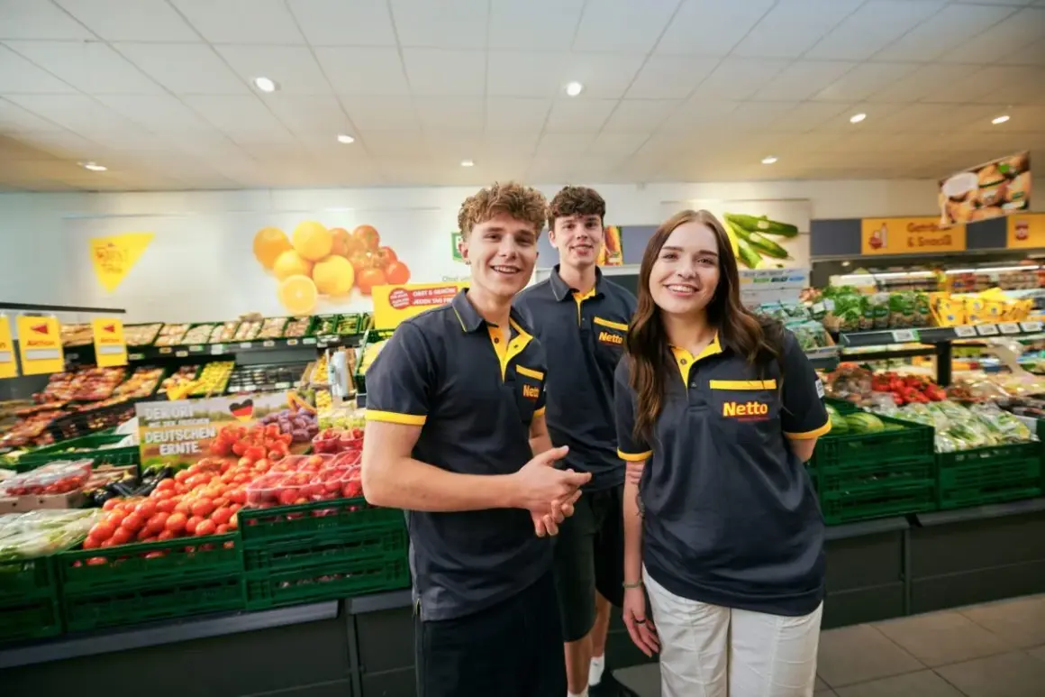 Drei junge Mitarbeiter lächeln in einem Supermarkt. Sie tragen Arbeitskleidung mit dem Netto-Logo. Die Ausbildung zum Elektroniker bietet ein gutes Gehalt netto.