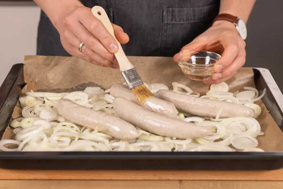 Bratwurst im Backofen: Zwiebelringe auf Backblech, darauf vier rohe Bratwürste. Mit Pinsel wird Öl aufgetragen.