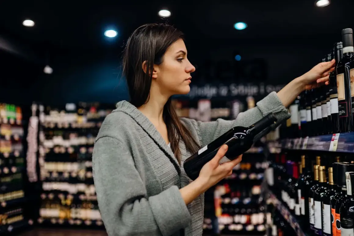 Donna sceglie una bottiglia di vino, pensando ai solfiti. Scaffali pieni di bottiglie sullo sfondo.