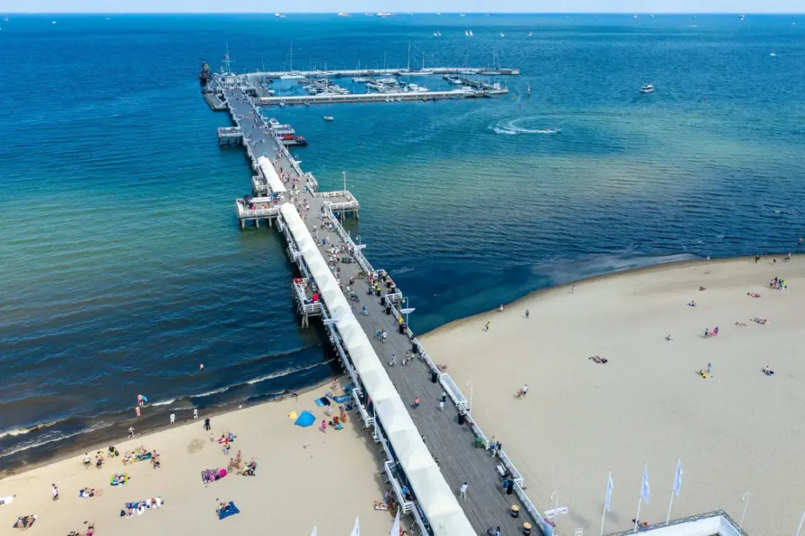 Słoneczny dzień nad Bałtykiem. Molo w Polsce tętni życiem, spacerują po nim tłumy ludzi. Na plaży odpoczywają plażowicze, a w oddali widać jachty.