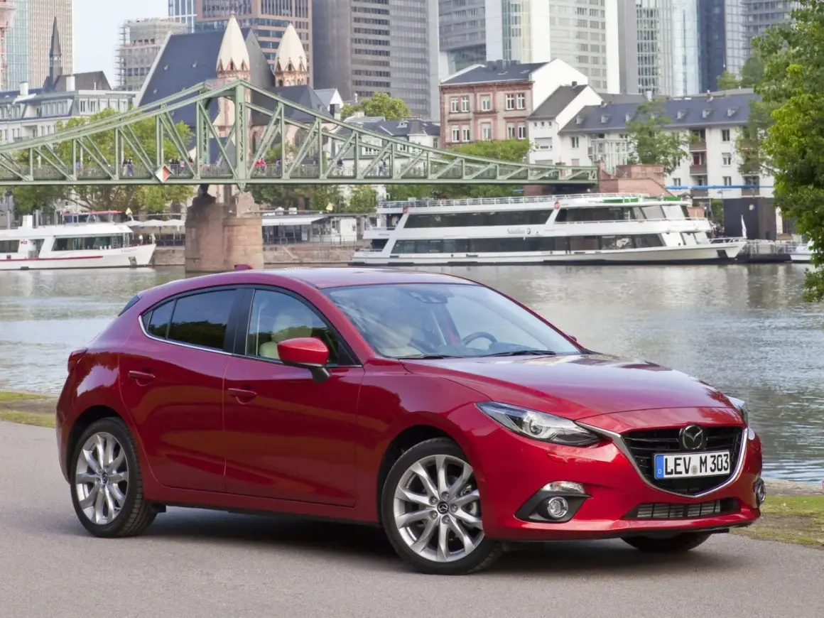 Czerwona Mazda 3 na tle miasta i rzeki. Wiele osób szuka takich aut, bo mazda 3 opinie ma zazwyczaj bardzo dobre.