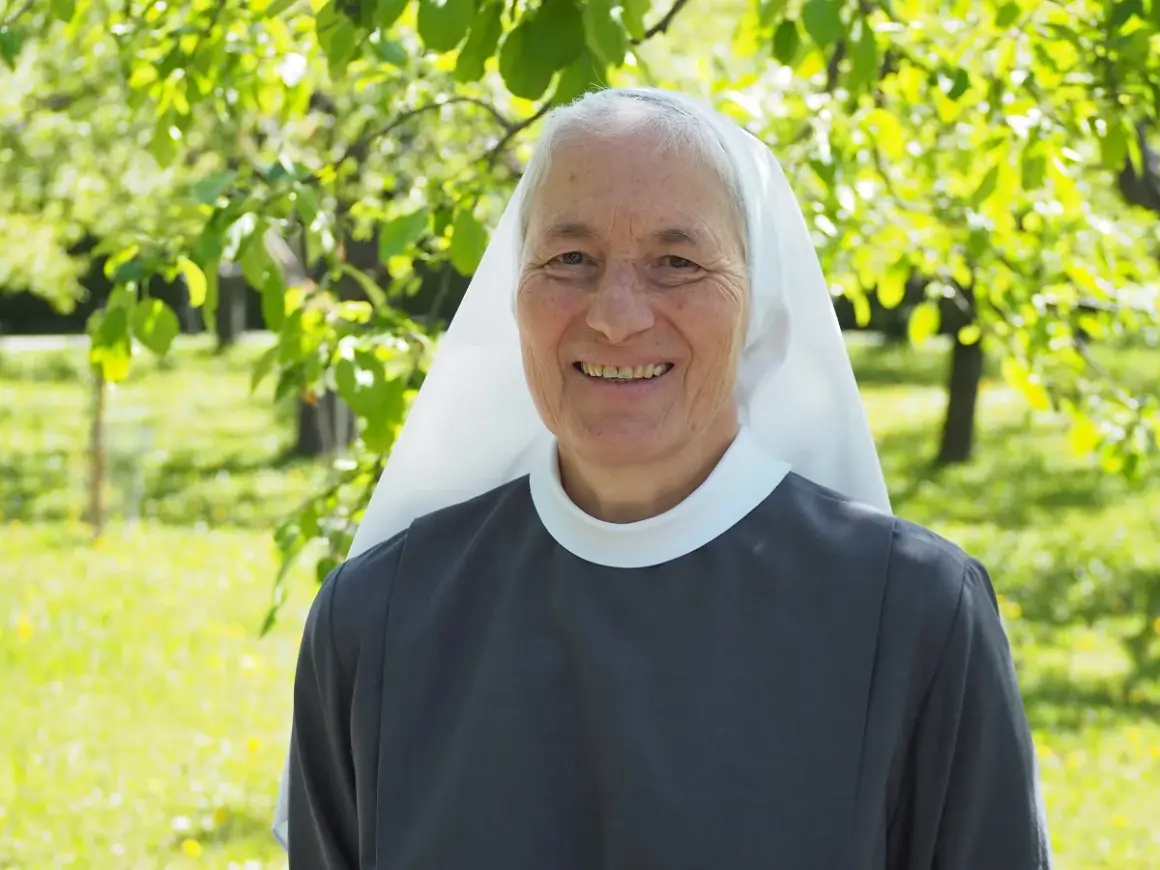 Lächelnde Nonne im Garten. Sie hat graue Haare und trägt eine graue Ordenskleidung mit weißem Schleier. Das Leben ist nicht fair, aber sie strahlt Hoffnung aus.