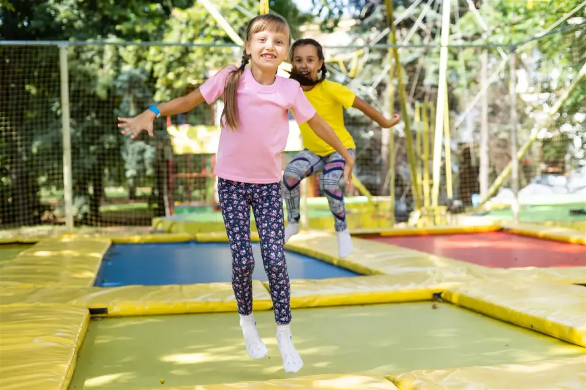 Dwie dziewczynki skaczą na trampolinie, śmiejąc się radośnie. Czy trampolina jest zdrowa dla dziewczynek? Z pewnością tak, to świetna zabawa i aktywność fizyczna!
