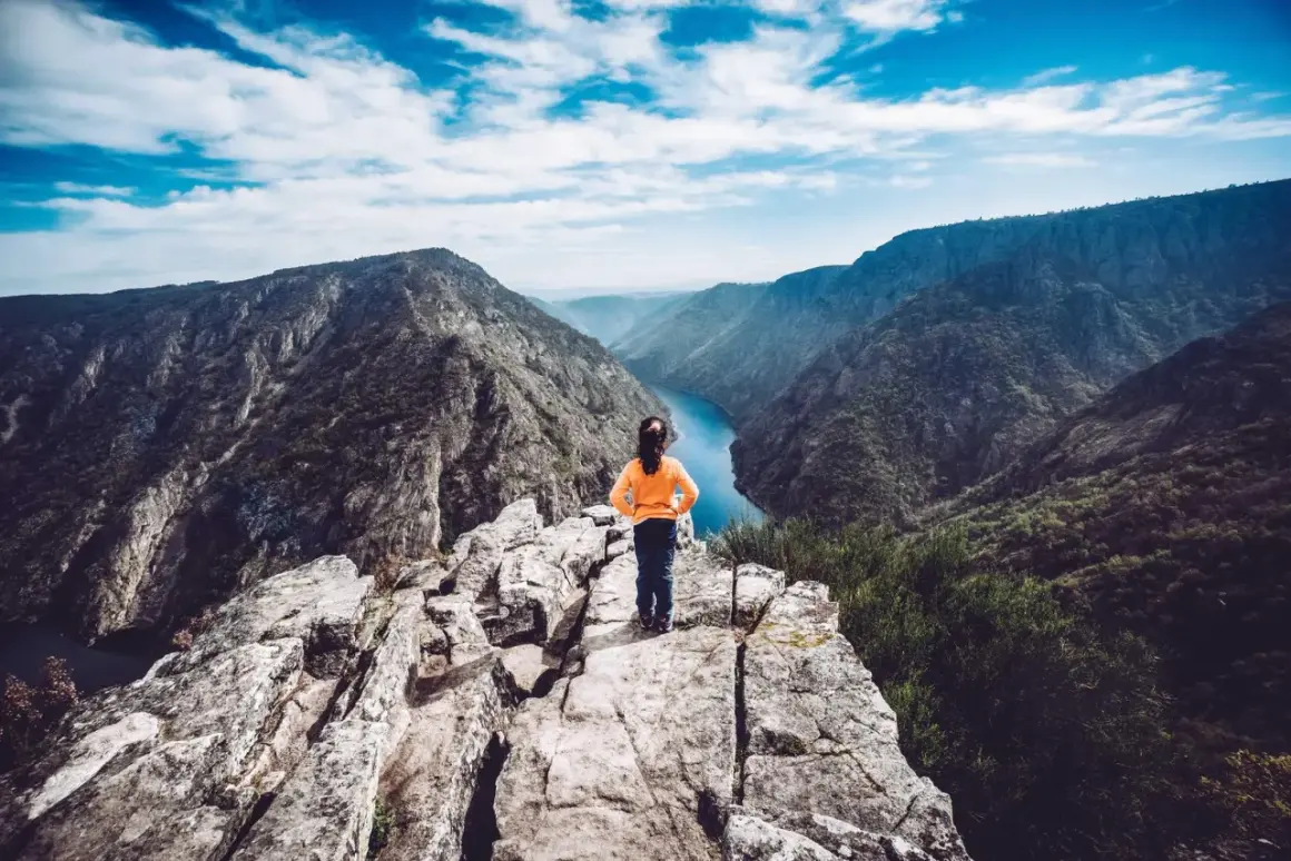 Niña en el mirador Vilouxe, contemplando un cañón con un río azul y montañas verdes bajo un cielo nublado.