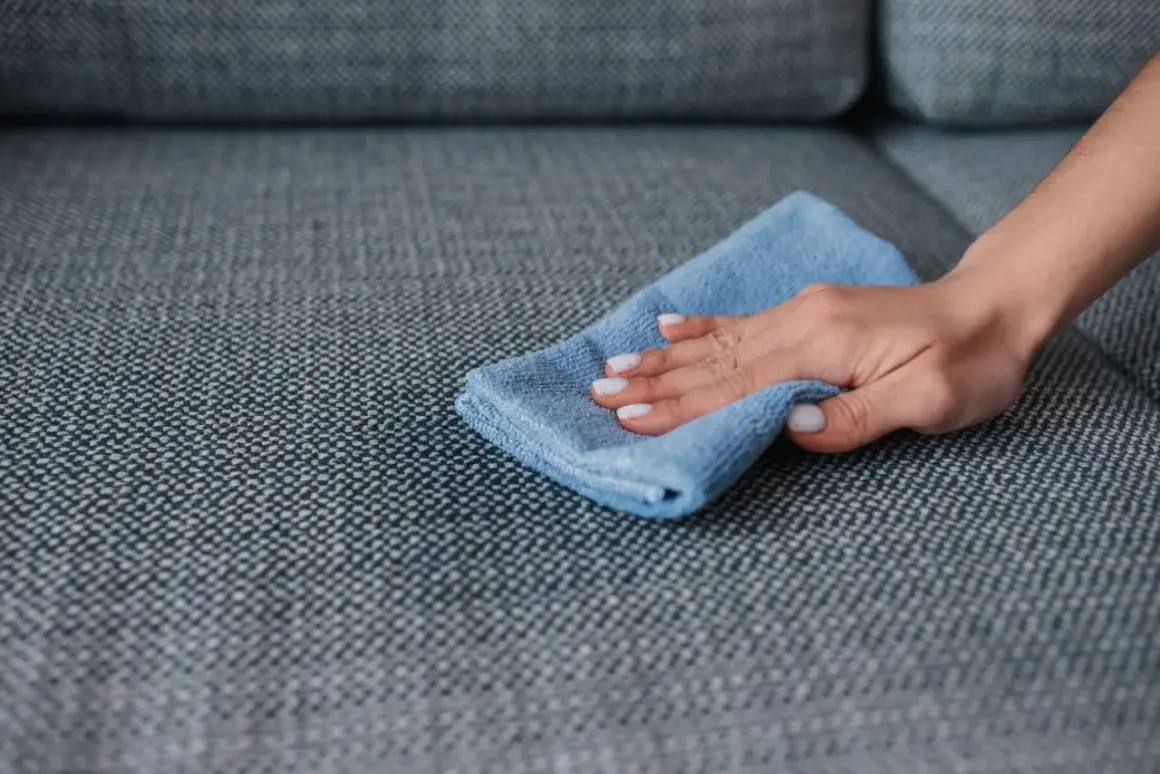 A hand with white nail polish wipes a grey couch with a blue microfiber cloth, demonstrating how to remove slime from couch.