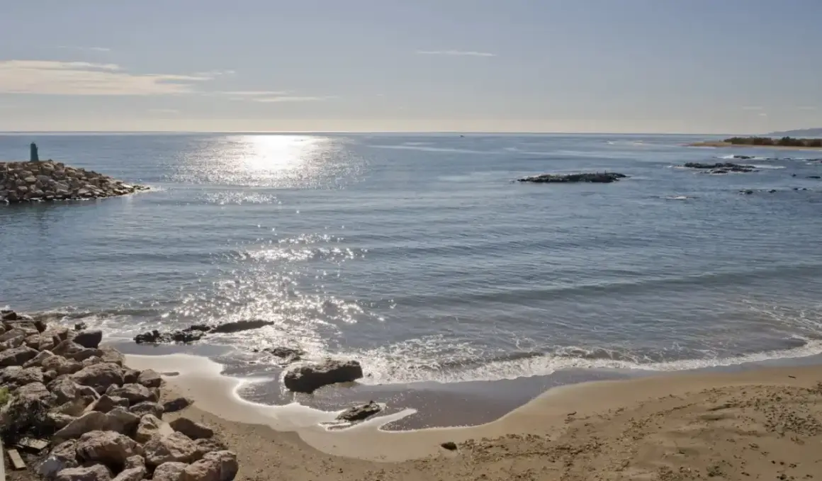 Costa de Villaricos con un faro en un rompeolas de rocas. El sol brilla sobre el mar azul y la arena dorada.