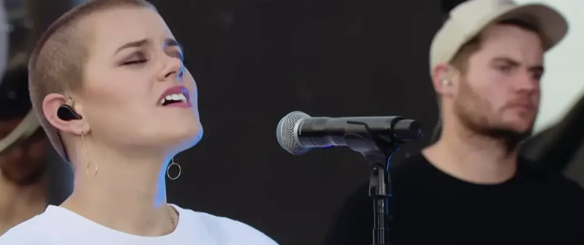 Taya Smith singt mit geschlossenen Augen und In-Ear-Monitoring. Ein Mann mit Kappe steht im Hintergrund.