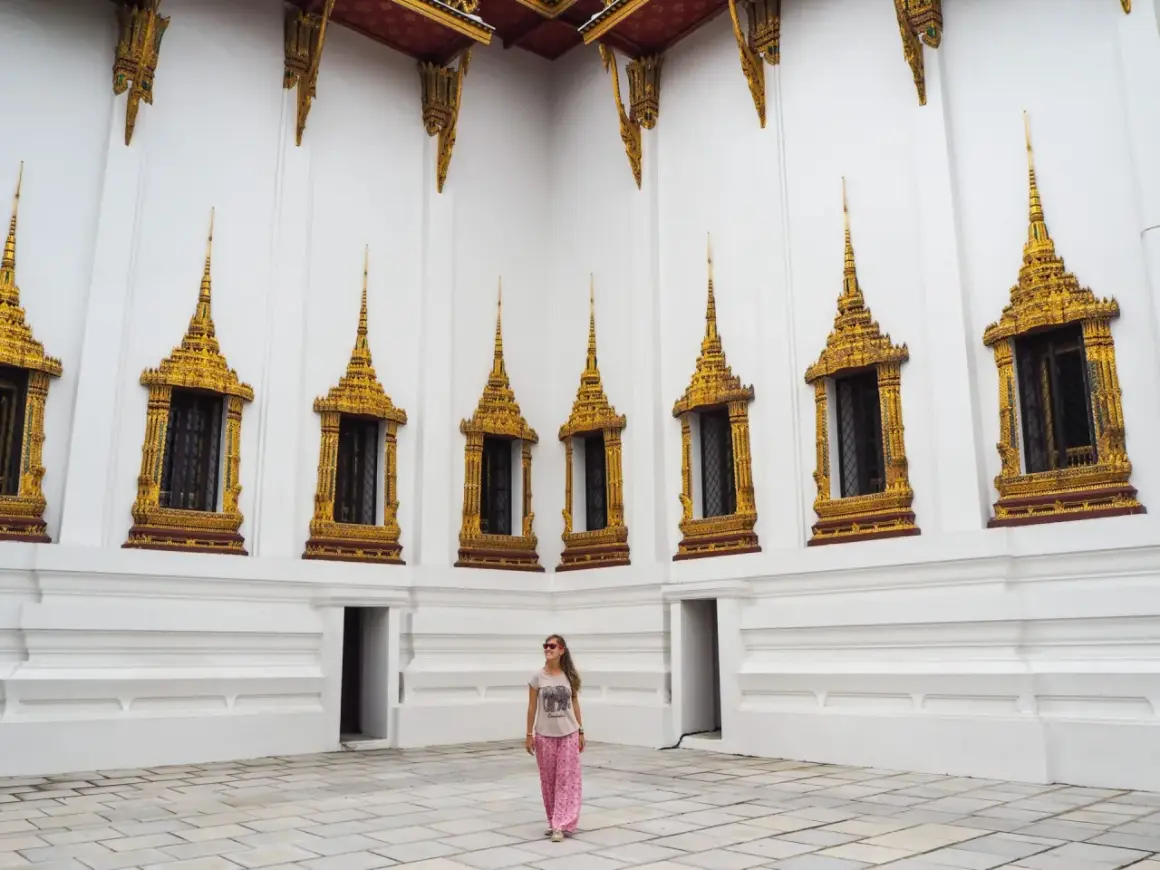 Una mujer sonríe frente a una pared blanca con ventanas doradas ornamentadas, un ejemplo de qué ver en Tailandia.
