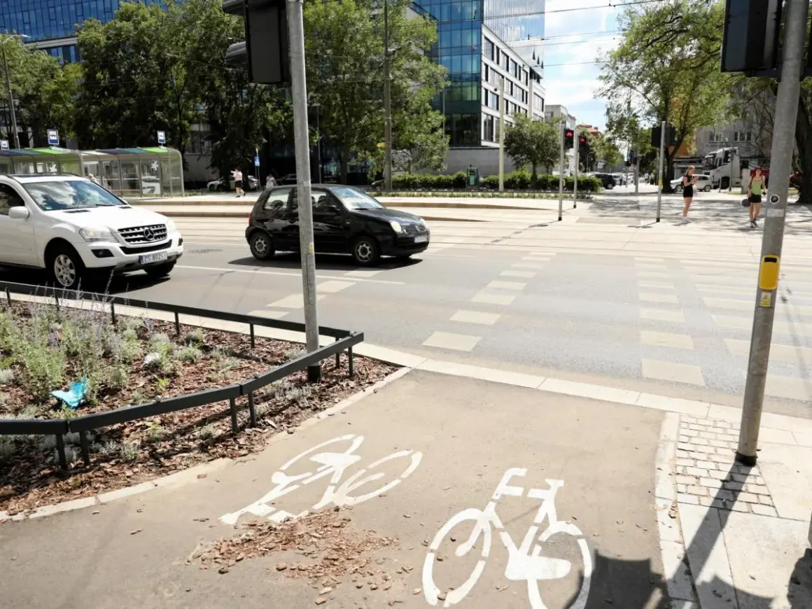 Czarny samochód wyprzedza na pasach, omijając białe auto. Na ścieżce rowerowej widoczne są symbole rowerów.