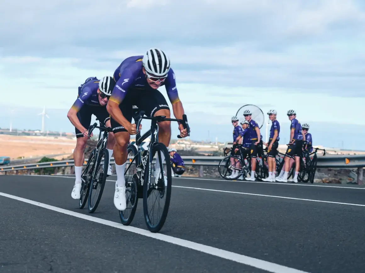 Cykla på Gran Canaria i tävlingsform. Ett cykellag i lila dräkter susar fram på en asfalterad väg med vindkraftverk i bakgrunden.