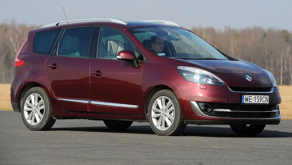 Ciemnoczerwony Renault Scenic 3 z kierowcą za kierownicą jedzie po drodze, w tle drzewa.