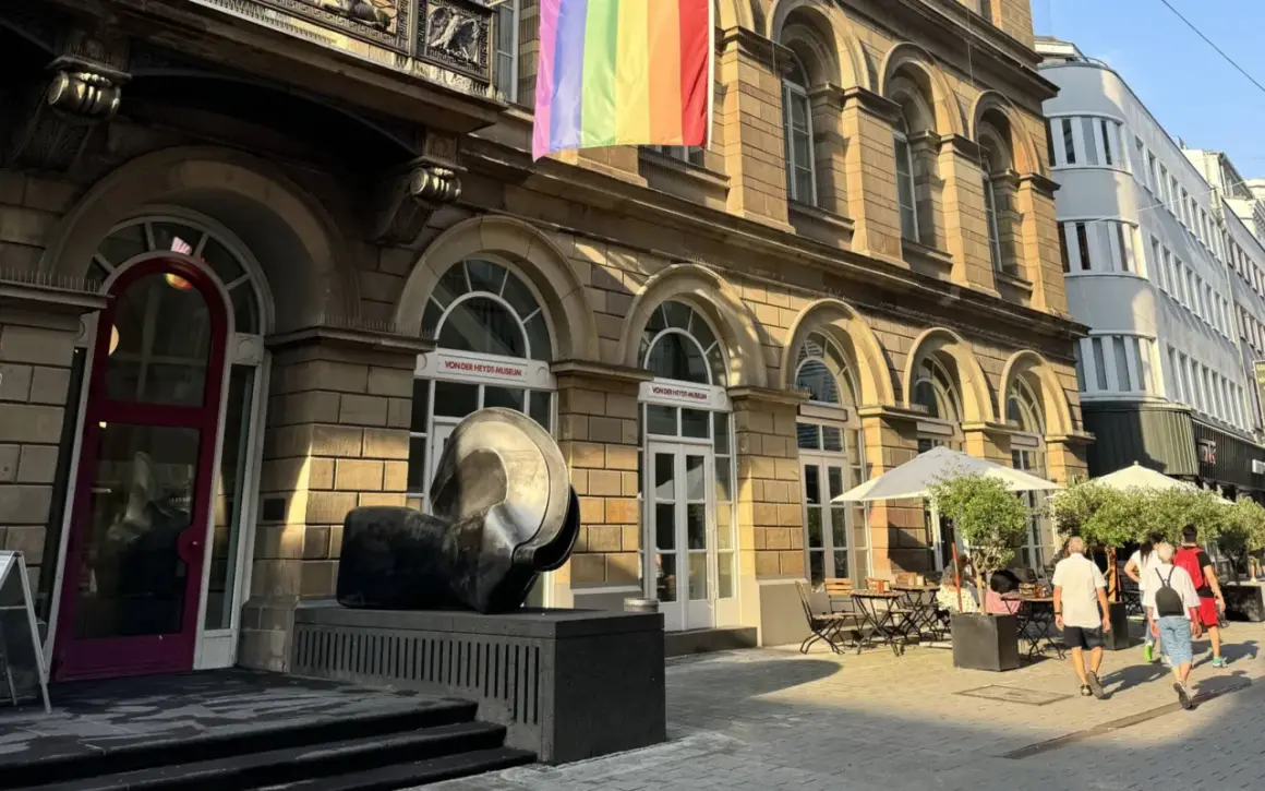 Ein Regenbogenbanner weht über dem von der Heydt Museum. Eine Skulptur steht davor, daneben ein Café mit Sonnenschirmen.