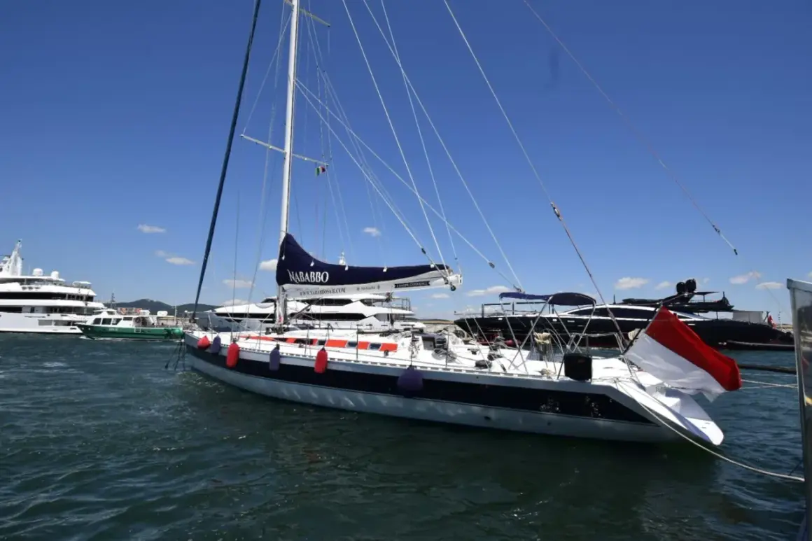 Barca a vela "Nababbo" con bandiera rossa e bianca, in porto con altre imbarcazioni. Sembra una cometa 12.