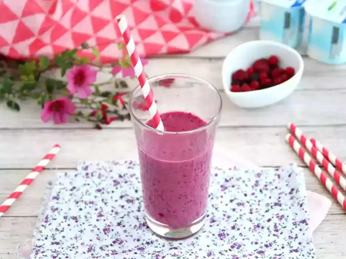 Ett glas med en lila indisk yoghurtdryck, dekorerad med ett rött och vitt randigt sugrör. Bredvid finns en skål med bär och yoghurtburkar.
