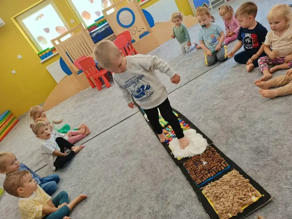 Dzieci aktywnie w żłobku ćwiczą zmysły na ścieżce sensorycznej. Maluchy z zaciekawieniem obserwują kolegę stąpającego po różnych fakturach.