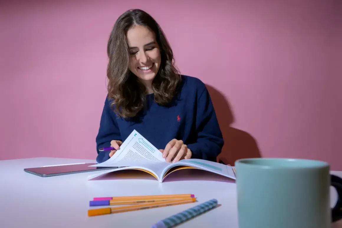 Lächelnde Studentin blättert in einem Buch, während sie lernt. Das Psychologiestudium ist schwer, aber sie meistert es. Bunte Stifte liegen bereit.