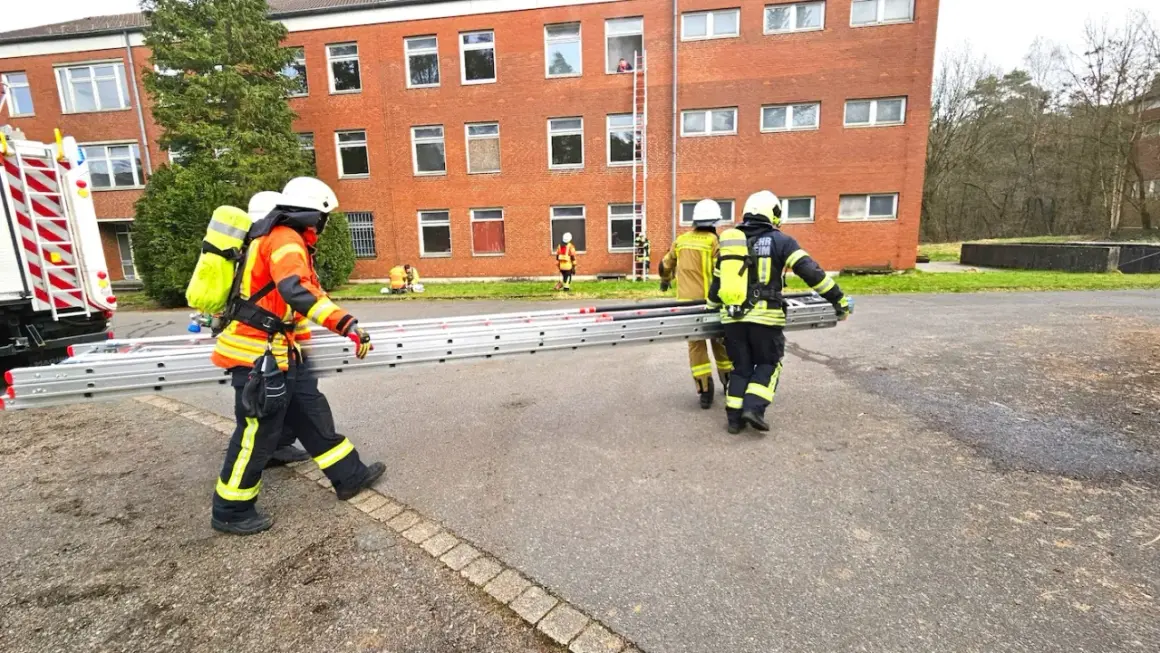 Feuerwehr-Messe: Einsatzkräfte transportieren eine Leiter vor einem Backsteingebäude. Eine Person steigt bereits eine Leiter zum Fenster empor.