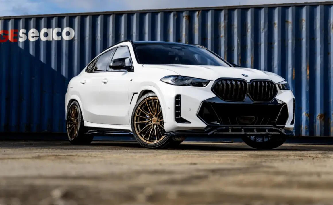 Białe BMW X6 z brązowymi felgami, na których zamontowano opony Imperial, prezentuje się imponująco na tle niebieskiego kontenera.