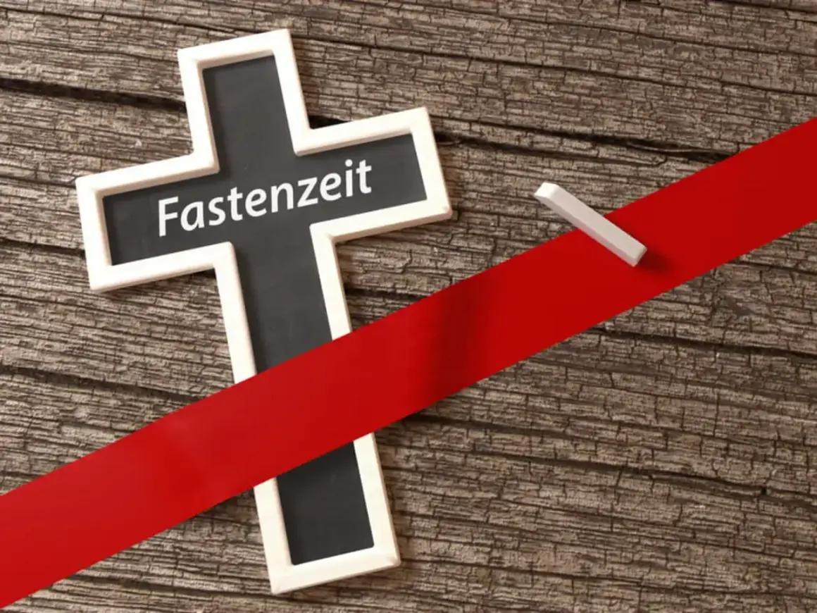 Kreuz mit der Aufschrift "Fastenzeit" auf Holzuntergrund, durchzogen von einem roten Band. Ein Stück Kreide liegt daneben.
