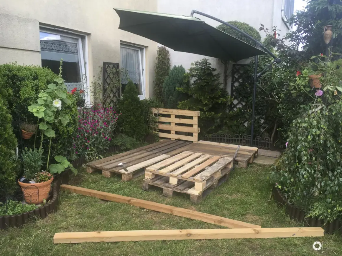 Ogród z meblami z palet. Podłoga z palet w ogrodzie czeka na wykończenie. Obok zielony parasol i roślinność.