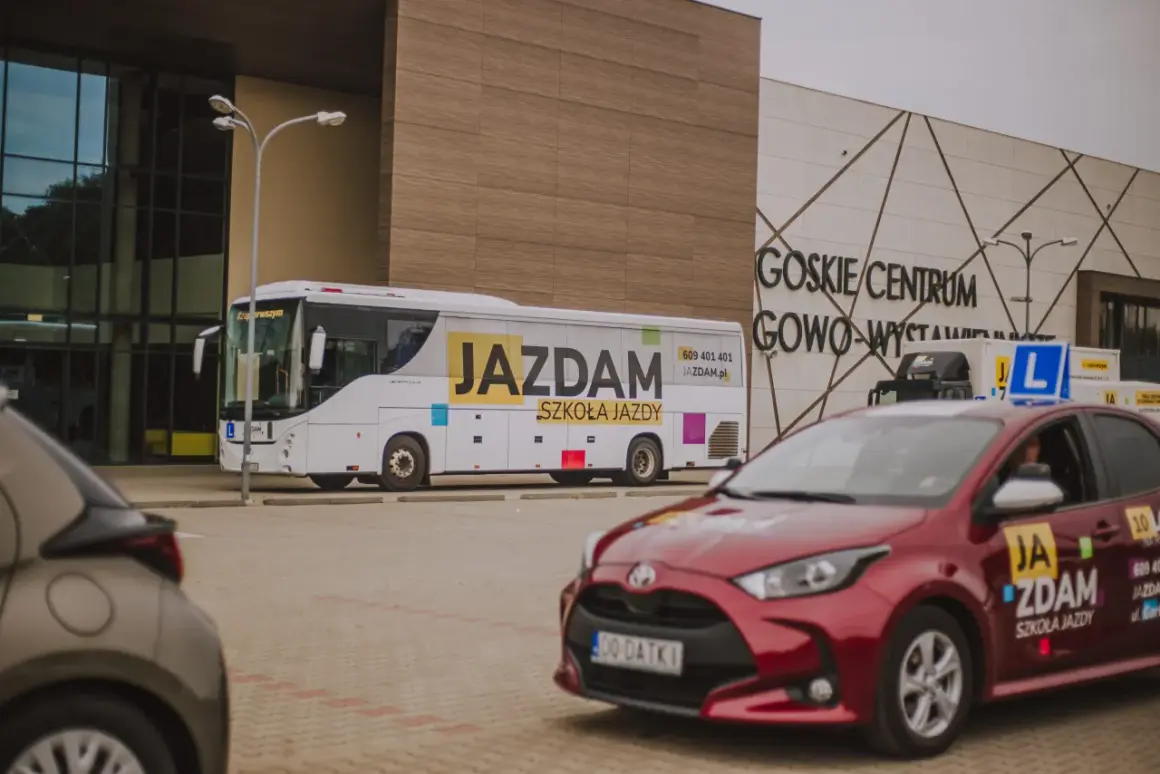 Autobus i czerwony samochód z napisem "JAZDAM SZKOŁA JAZDY" przed budynkiem GOSKIE CENTRUM GOWO-WYSTAWIENNICZE.